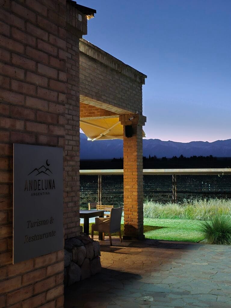 Fachada de la Bodega Andeluna en Mendoza al atardecer, con cartel de Turismo y Restaurante en primer plano, galería exterior, viñedos y la silueta de la cordillera de los Andes de fondo.