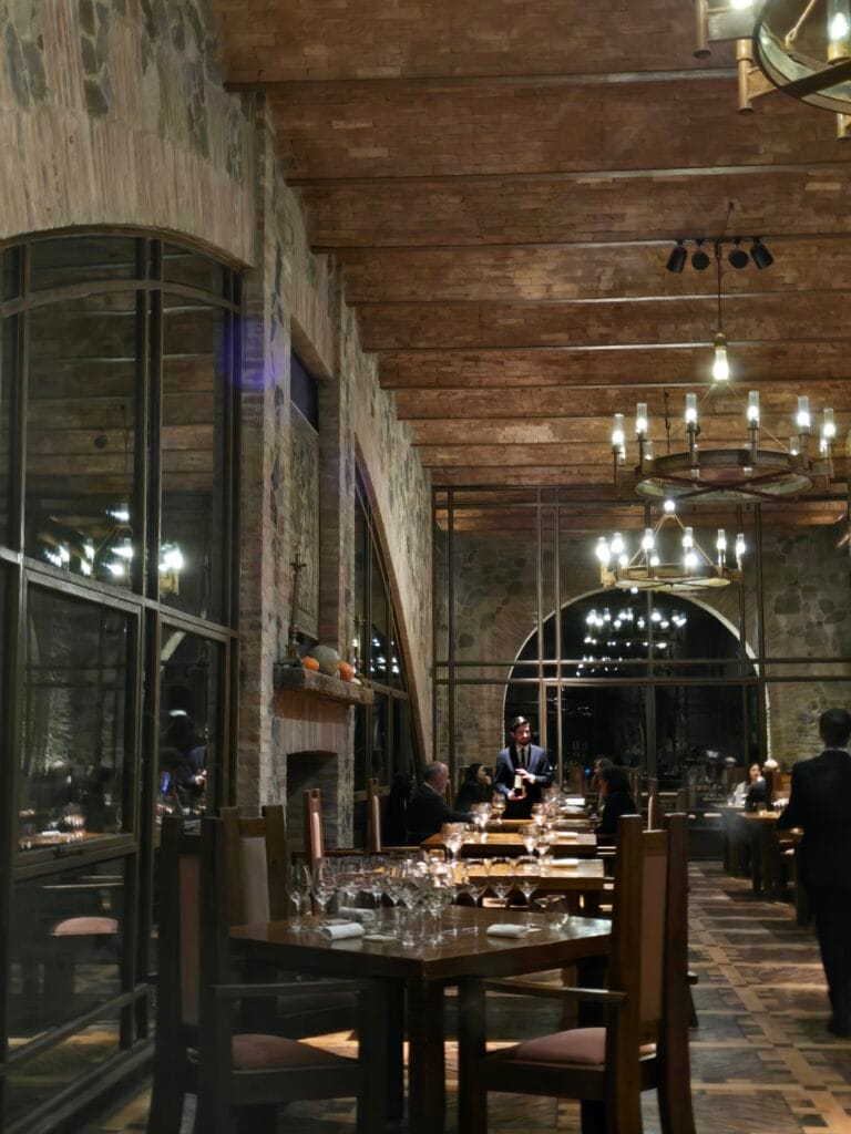 Interior del restaurante Angélica Cocina de Viñedo durante el servicio de cenas en Mendoza, con mesas preparadas con copas de vino, lámparas de hierro encendidas y comensales interactuando con un mozo.