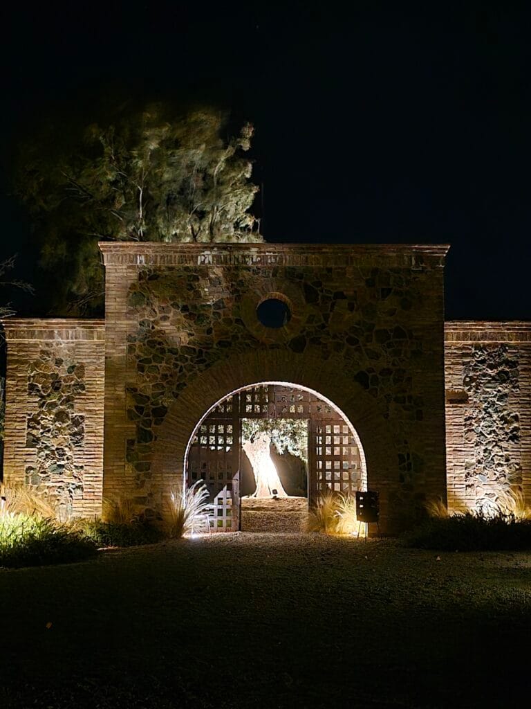 Fachada iluminada de noche en Angélica Cocina de Viñedo para la propuesta de cenas en Mendoza, con un gran arco central de piedra y ladrillo.