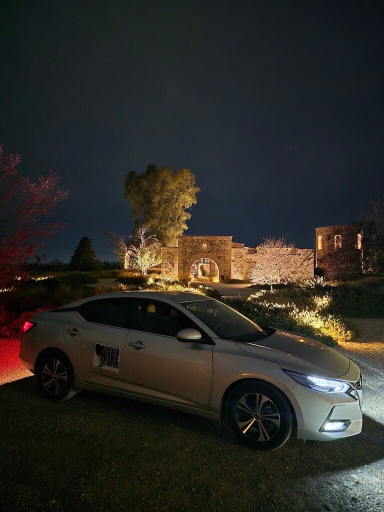 Vehículo privado de Mendoza Wine Transfers estacionado de noche frente a la entrada iluminada de piedra de Angélica Cocina de Viñedo para servicio de cenas.